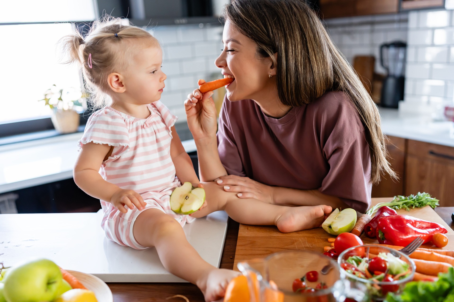 Cheerful mother and her cute little baby are eating healthy fruit and vegetables in the home kitchen